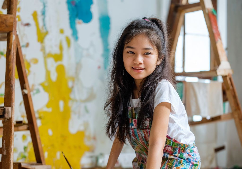 カラフルなアートスタジオで笑顔の少女アーティスト。背景に黄色とターコイズの抽象的な壁画。
